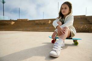 Portrait of adorable little girl looking into the distance while sitting on her skateboard outdoors. photo