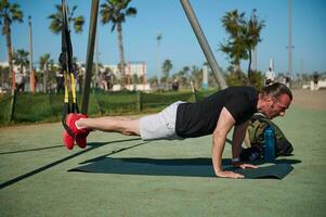 Caucasian young male athlete doing push ups, putting feet inside suspension fitness straps while working out outdoors photo