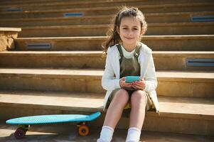 Authentic little girl, elementary school student holding mobile phone, smiles cutely looking confidently at camera photo