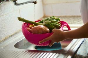 de cerca mujer ama de casa Lavado Fresco vegetales debajo corriendo agua, en pie por inoxidable lavabo en el cocina a hogar foto