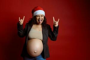 Charismatic emotional pregnant woman grimacing, standing isolated over red color background, dressed in Santa hat photo