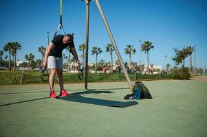 Full length portrait of strong muscular athlete performing bodyweight training with suspension straps outdoors photo