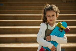 Authentic charming Caucasian kid girl 6 years old, holding skateboard outdoors, cutely smiles looking at camera photo