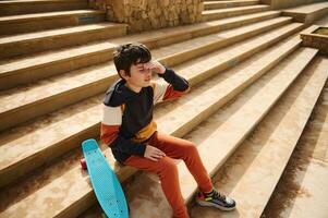 Adolescente estudiante chico, colegial en ropa de deporte sentado en pasos en el urbano Parque de patinaje, pensativamente mirando dentro distancia foto