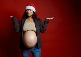 carismático embarazada mujer, expectante madre en elegante gris chaqueta de sport y Papa Noel sombrero, participación Copiar espacio en manos palmas arriba foto