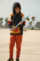 Caucasian teenage boy dressed in stylish sportswear, holding skateboard and looking down, standing at outdoor skatepark photo