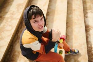 Handsome teenager boy holding skateboard and dreamily looking at camera, sitting on steps outdoors. People and lifestyle photo