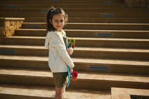 caucásico contento sonriente elemental años colegio niño patinador con patineta exterior. activo pequeño niña con centavo tablero foto