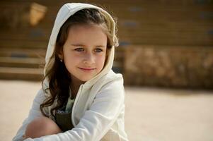 Close-up little girl with hood on her head, smiling cutely and dreamily looking away, sitting on a skateboard outdoor photo