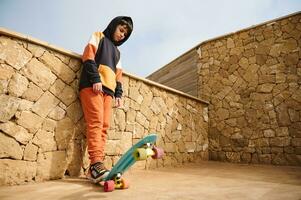 Handsome teen boy in sportswear, playing with a skateboard, standing with hands in pockets against stone wall background photo