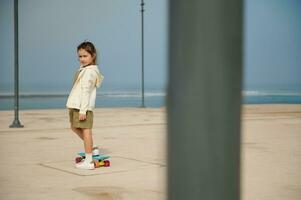 adorable niño niña mirando a cámara, en pie en patineta en uno pierna a patinar parque, en contra Oceano antecedentes foto