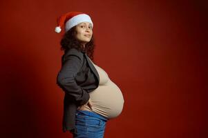 Side portrait pretty pregnant woman in Santa hat, showing her big belly in pregnancy third trimester, smiles at camera photo