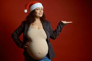 atractivo embarazada mujer vistiendo rojo Papa Noel sombrero, sonrisas mirando a cámara, sostiene un Copiar anuncio espacio en su mano palma arriba foto