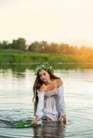Beautiful black haired girl in white vintage dress and wreath of flowers standing in water of lake. Sun flare. photo