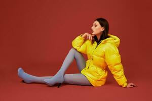 Studio portrait of young brunette woman sitting on the floor in yellow down jacket and grey blue panty hoses or stockings photo