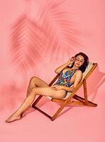 Summer lifestyle fashion portrait of young stunning woman on a lounge chair, pink background, studio shot. Wearing stylish sunglasses, bikini. photo