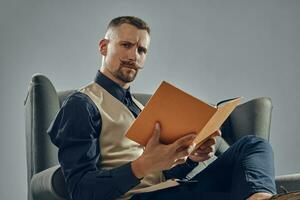 hombre con elegante Bigote, vestido en negro camisa y pantalones, beige chaleco es sentado en oscuro sofá, leyendo un revista. gris fondo, de cerca. foto