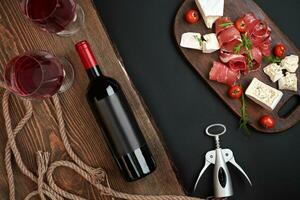 Above overhead view flat lay still life of assortment various cheese and delicatessen and red wine on a old wooden board on black table photo