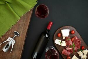 Above overhead view flat lay still life of assortment various cheese and delicatessen and red wine on a old wooden board on black table photo