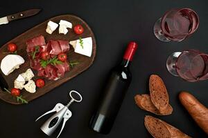 Red wine, cheese, cherry tomato, bread and prosciutto on wooden board over black backdrop, top view, copy space photo