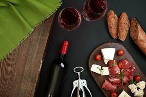 Above overhead view flat lay still life of assortment various cheese and delicatessen with traditional bread and red wine on a old wooden board on black table photo