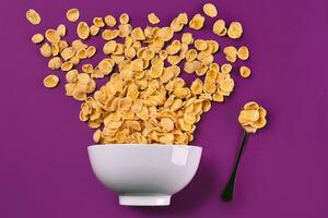 Bowl with corn flakes and spoon on purple background, top view photo