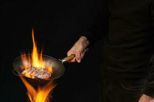 Young fellow dressed in jumper and jeans. He is holding burning wok pan above fire and frying beef steak, against black studio background. Side view photo