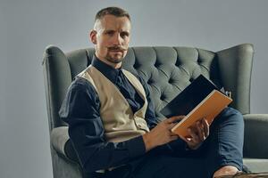 hombre con elegante Bigote, vestido en negro camisa y pantalones, beige chaleco es sentado en oscuro sofá, leyendo un revista. gris fondo, de cerca. foto