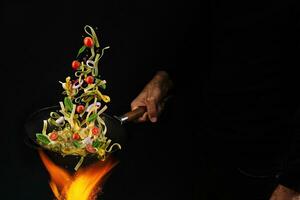 Unrecognizable man holding wok pan above fire and cooking pasta with cherry tomatoes, onion and basil against black background. Close up photo