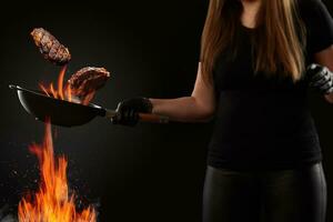 Cook with tattooed hands, dressed in leggings and t-shirt. Holding wok pan above fire and frying two beef steaks against black background. Side view photo