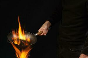 Young guy dressed in jumper and jeans. He is holding burning wok pan above fire, against black studio background. Cooking concept. Close up photo