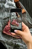 Photographing food concept - woman takes picture of raw dry aged t-bone steaks for grill with fresh herbs and cleaver photo