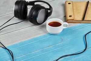 Black headphones with cord, brown notebook in leather cover, pencil and cup of tea on blue and white wooden tabletop. Close up, copy space photo