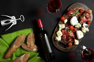 Delicious appetizer to wine - ham, cheese, baguette slices, tomatoes, served on a wooden board, and glass with red wine on black surface photo