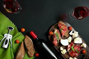 Prosciutto, salami, baguette slices, tomatoes and nutson rustic wooden board, two glasses of red wine over black background photo