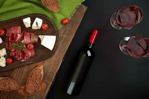 Red wine, cheese, cherry tomato, bread and prosciutto on wooden board over black backdrop, top view, copy space photo