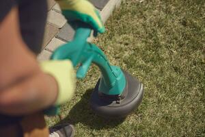 Gardener in sneakers and colorful gloves is mowing green grass with handheld modern lawn mower in courtyard, near a tiled path. Close up, top view photo