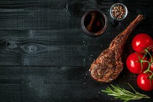 parilla seco Envejecido costilla de carne de vaca con especia, vegetales y vaso de rojo vino de cerca en negro de madera antecedentes foto