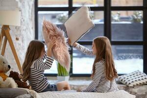 The image of two little sisters sitting on the bed in the room. They using pillow to fight each other photo