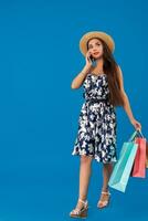 Shopping concept. Beautiful smiling brunette with shopping bags talking on the phone on blue studio background with copy space. photo