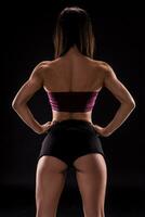 Young beautiful dark-haired woman in dark purple sports bra and black short shorts seductively posing with her back against a black background photo