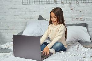 Cute little girl girl feeling amusing while watching cartoons on a laptop sitting on bed photo