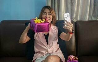 woman sitting on sofa calling via video with smartphone to friend showing gift box and smiling happiness long distance relationship photo