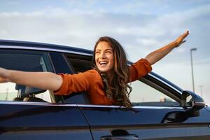 joven hermosa mujer consiguiendo nuevo coche. ella muy contento y entusiasmado. sonriente hembra conducción vehículo en el la carretera en un brillante día. pega su cabeza fuera de el parabrisas foto