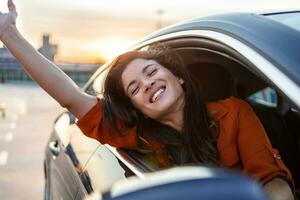 Young woman sitting in a car hand out of window. Happy woman driving a car and smiling. Portrait of happy female driver steering car with safety belt. Cute young lady happy driving car. photo