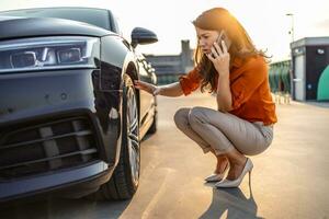 mujer vocación para asistencia con plano neumático en coche en el ciudad. joven mujer con alto tacones en el ciudad con plano neumático, frustrado cerca roto coche foto