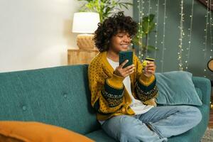 mujer afroamericana comprando en línea con un teléfono inteligente pagando con tarjeta de crédito dorada. niña sentada en casa comprando en internet ingrese los detalles de la tarjeta de crédito. servicio de entrega de comercio electrónico de compras en línea. foto