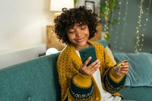 mujer afroamericana comprando en línea con un teléfono inteligente pagando con tarjeta de crédito dorada. niña sentada en casa comprando en internet ingrese los detalles de la tarjeta de crédito. servicio de entrega de comercio electrónico de compras en línea. foto