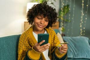 mujer afroamericana comprando en línea con un teléfono inteligente pagando con tarjeta de crédito dorada. niña sentada en casa comprando en internet ingrese los detalles de la tarjeta de crédito. servicio de entrega de comercio electrónico de compras en línea. foto