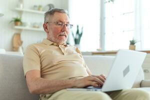 Confident stylish happy middle aged senior man using laptop at home. Stylish older mature 60s beard grandfather sitting at couch looking at computer screen typing chatting reading writing email. photo